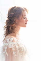 Beautiful natural redhead girl bride, with nude makeup, wearing a white dress, stands at the window next to a transparent curtain.