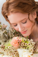 Fototapeta premium Beautiful natural redhead girl bride, with nude makeup, wearing a white dress, tenderly holds a wedding bouquet in her hands. Close up portrait.
