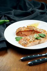 Serving appetizing salmon steak on a white plate in a composition with a black cloth and ingredients on a wooden table. Grilled seafood