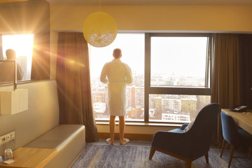 Silhouette of a young man with cup of coffee in the white bathrobe on the hotel window background at the yellow sunrise time