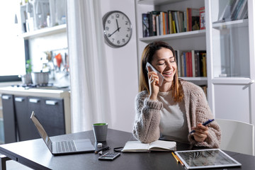 Giovane donna pratica lo smart working da casa, e parla al telefono sorridendo , sullo sfondo la...