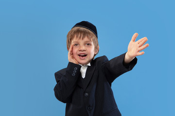 Calling, shouting. Portrait of a young orthodox jewish boy isolated on blue studio background. Purim, business, festival, holiday, childhood, celebration Pesach or Passover, judaism, religion concept.