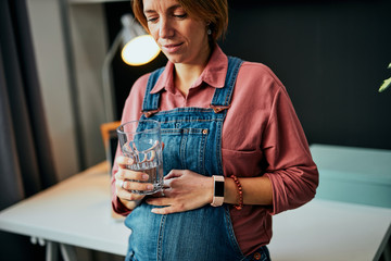 Adorable Caucasian pregnant freelancer standing in her office, touching belly, drinking water and taking a break from work. Work from home concept.