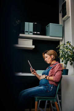 Side view of cute Caucasian pregnant woman sitting in home office and using tablet. Work from home concept.