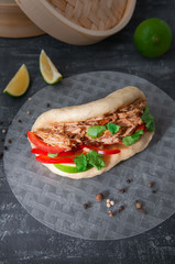 Bao burger with baked pork, sweet pepper, cilantro and lime. Gua bao is a traditional asian steamed bun. Lies on rice paper. The background is decorated with spices, herbs and lime. View from above.