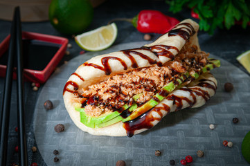 Bao burger with salmon, avocado and balsamic dressing. Gua bao traditional asian steamed bun. Lies on rice paper. The background is decorated with spices, herbs and lime. Close-up.