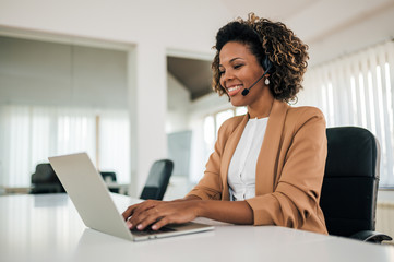 Smiling latin businesswoman working with laptop and headset, portrait. Cheerful assistant in the bright office.