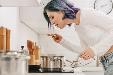 Selective focus of girl with colorful hair preparing food and holding pan lid with spatula near kitchen stove
