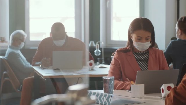 Mixed raced woman in protective face mask sitting at desk and typing on laptop while working in open space office with colleagues during coronavirus outbreak