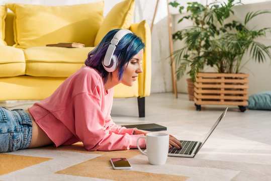 Freelancer with colorful hair and headphones working with laptop near cup of tea and smartphone on floor in living room