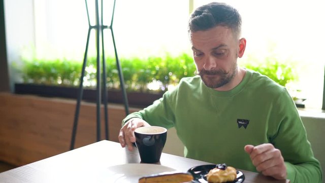 Bearded man with curled up mustache eats chocolate eclair and drinks coffee in cafe