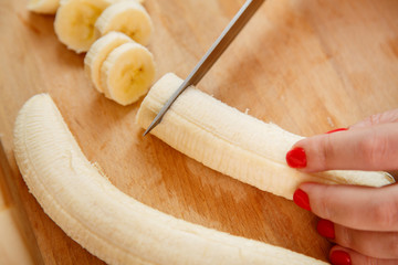 Female hand cuts banana with smoothie rings on a wooden board with a knife.