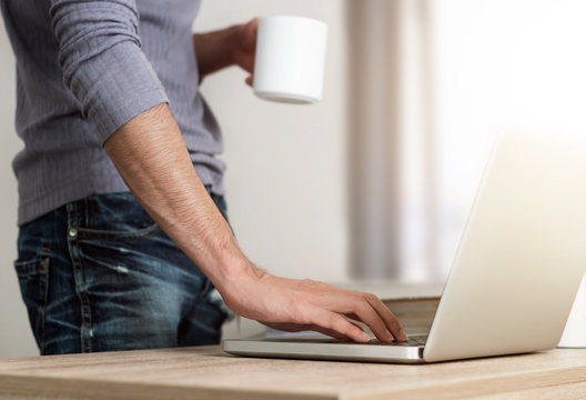 Close up low angle view of a man standing at the a desk surfing on internet and drinking coffee in morning.