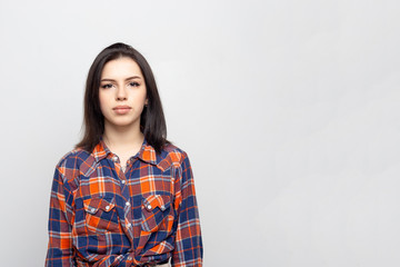 Fototapeta premium Portrait young beautiful woman not smiling in plaid shirt isolated on white-gray background. Girl with minimal natural make-up on her face. The photo is not retouched with natural defects on the skin.