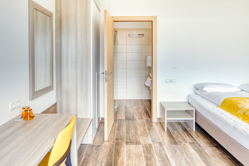 Modern minimalist hotel room with open bathroom door, mirror, glasses, yellow chair and wardrobe. Wooden tile floor. Plain white wall without decoration.