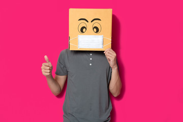 Man wearing cardboard box with mask to protect from COVID 19 virus showing thumbs up sign against pink background