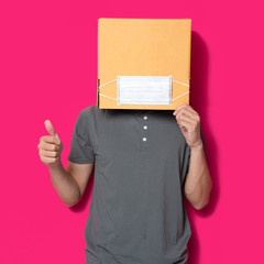 Man wearing cardboard box with mask to protect from COVID 19 virus showing thumbs up sign against pink background