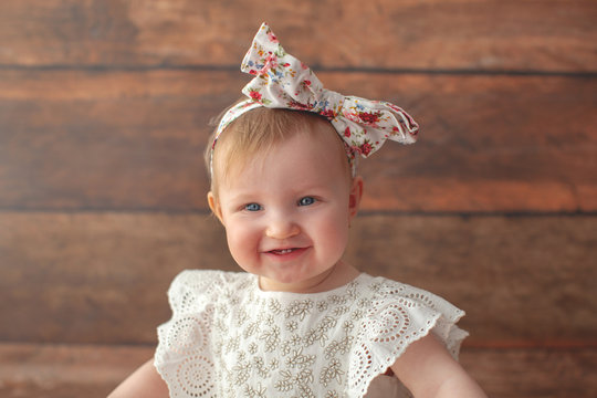 Portrait of the cute one year old girl. With headband and in white dress. On wooden background. Caucasian toddler little baby. With first teeth. 