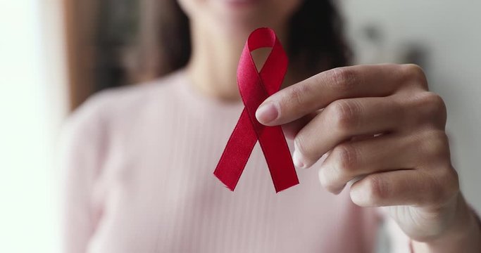 Young woman holds red ribbon in hand showing to camera. HIV awareness, AIDs treatment prevention and protection, volunteer support in charity, donation. World breast cancer day concept. Close up view