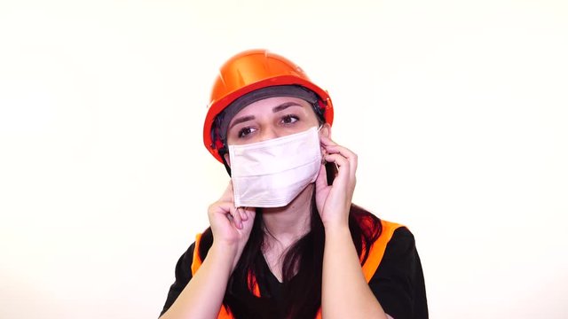 Female construction worker in overalls putting on medical mask on face on white background.