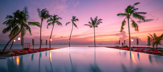 Beautiful poolside and sunset sky with palm trees silhouette. Luxurious tropical beach landscape, deck chairs and loungers and water reflection. Tranquil summer vacation, travel concept infinity pool - Powered by Adobe