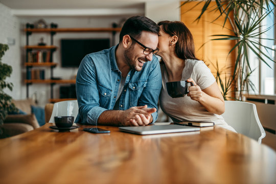 Portrait of a couple having fun at home. Woman whispering to man's ear, he is laughing. - Powered by Adobe