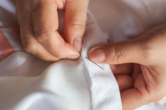 Hand of the seamstress is using a needle and white thread to sew the seam on the shirt close-up.