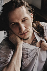 A guy with long black hair in jeans and a white shirt sits in a bathtub filled with water, next to the window