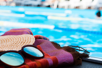 Vintage summer wicker straw beach hat, sun glasses and cover-up beachwear wrap near swimming pool, tropical background.