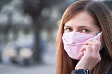 Young woman with pink cotton hand made face nose mouth mask talking over her mobile phone, close portrait. Can be used during coronavirus covid-19 outbreak prevention