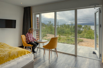 Teleworking. Middle-aged man with curly blond hair, white-red checkered shirt. Sitting with a laptop at a table in a modern hotel room next to his bed. Country views on a gray day. Beautiful views