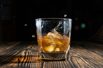Whiskey with ice cubes on a wooden table. An old tabletop with light and a glass of strong drink. A splash of whiskey in a glass while serving ice.