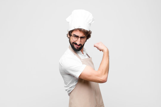 young crazy chef feeling happy, satisfied and powerful, flexing fit and muscular biceps, looking strong after the gym against white wall