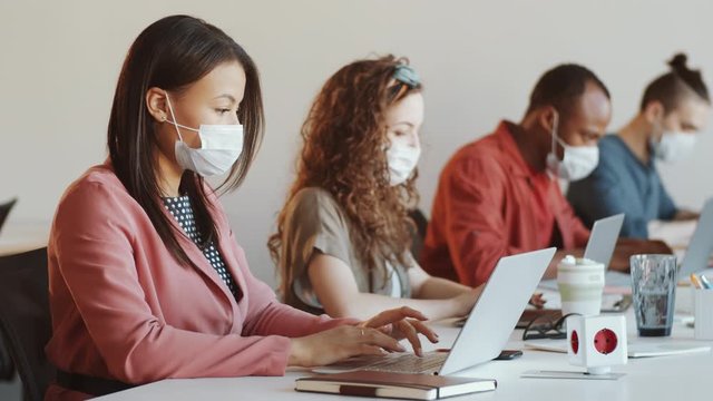 Young mixed raced businesswoman in face mask using laptop at desk while working in open space office with diverse colleagues during covid-19 outbreak
