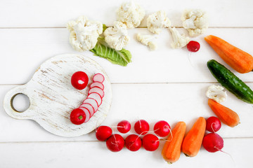 Fototapeta na wymiar Fresh vegetables on a white wooden table - cauliflower, radishes, cucumbers and carrots. On a round cutting Board sliced radishes