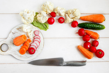 Fresh vegetables on a white wooden table - cauliflower, radishes, cucumbers and carrots. On a round cutting Board sliced radishes and carrot. Next to the vegetables is a large chef's knife