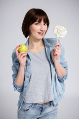 healthy and unhealthy food concept - portrait of young woman or teenage girl with braces on teeth holding lollipop and green apple over gray