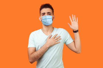 I promise. Portrait of serious brunette man with surgical medical mask standing with raised arm, swearing allegiance, taking oath with responsible expression. studio shot isolated, orange background