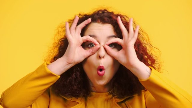cheerful girl imitating eyeglasses with hands and grimacing isolated on yellow