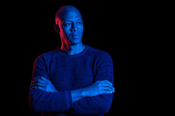 Black man with blue and red light, isolated on black background, looking sideways with arms crossed. Copyspace.