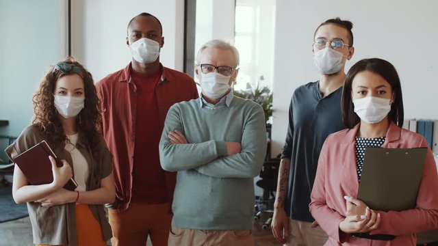 Business team of mixed-aged multiethnic men and women in protective face masks and casual clothes standing together in office and looking at camera while working during coronavirus outbreak