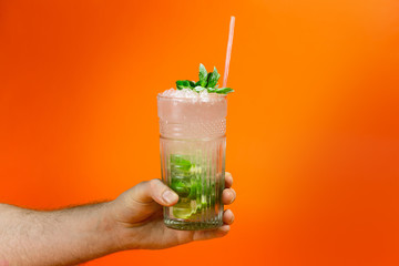 Close up of a man's hand holding a glass of tropical mojito cocktail on orange background with copy space. Summer time vacation concept.