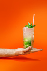 Close up of a man's hand holding a glass of tropical mojito cocktail on orange background with copy space. Summer time vacation concept.