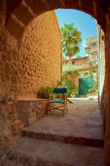 Pedestrian street in a small town. The street is made of stones and has some steps. There are stone houses, green doors and windows and a chair under a stone arcade. It is summer in Mallorca, Spain.