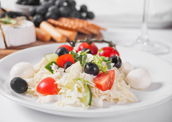 vegetables salad on the plate, glass of white wine and plate with assorted cheese, fruit and other snacks.