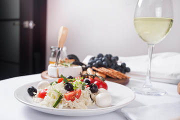 vegetables salad on the plate, glass of white wine and plate with assorted cheese, fruit and other snacks.