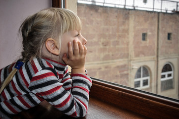 White-haired girl looks out the window. Quarantined child. The child is sad at the window.