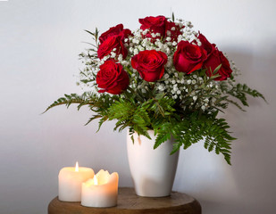 Beautiful large bouquet of dark red roses in a vase and two lighted candles on a small wooden table on a white background.