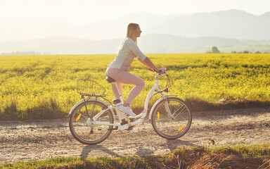 Fototapeta premium Young woman rides electric bicycle on dusty country road, view from side, sun backlight field with yellow flowers background