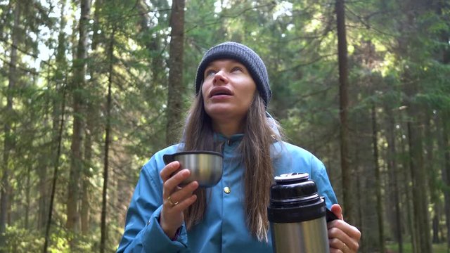 Woman in a blue raincoat and hat having rest with hot drink, tea from thermos sitting on the tree in the forest